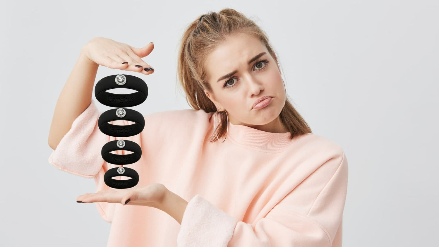 Young woman in a pink sweater holding and comparing stretched black Gemslique silicone rings with a puzzled facial expression.